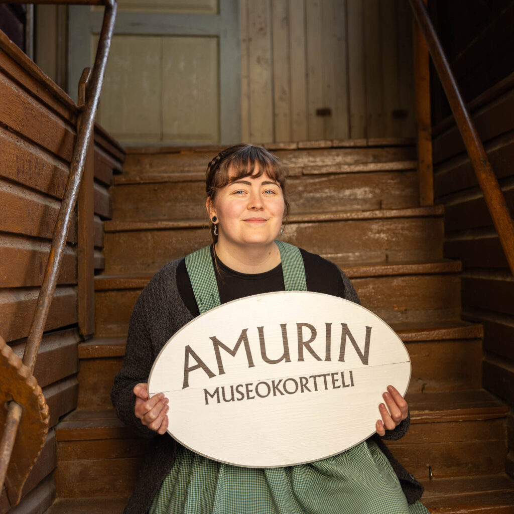 Tyttö vanhanaikaisissa vaatteissa istuu Amurin talon portailla ja pitelee käsissä kylttiä, jossa lukee Amurin museokortteli.