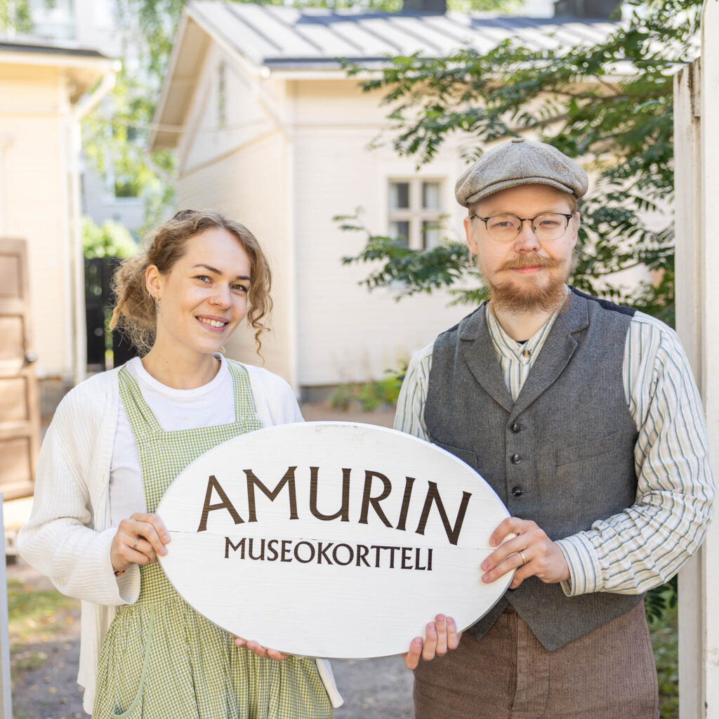 Two guides dressed in period costumes are holding a sign that reads "Amur Museum Quarter." The museum's wooden buildings are visible in the background.