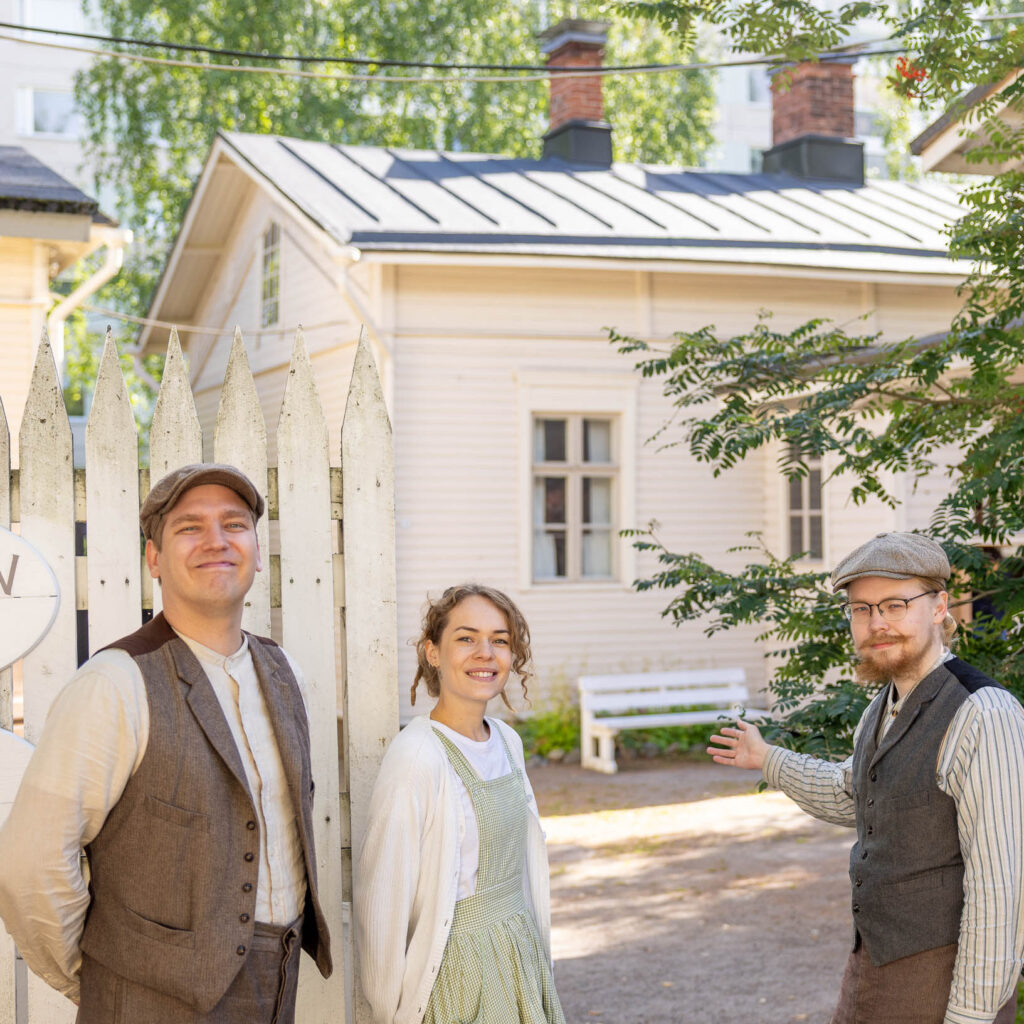Kolme vanhanaikaisiin vaatteisiin pukeutunutta henkilöä seisoo Amurin portilla, taustalla näkyy vanhoja puutaloja.